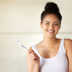 Brunette young woman in a white tanktop smiles while holding her toothbrush before her oral hygiene routine
